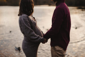 Couple maternity Photos in Autumn in London. This is a photo of a couple holding hands in front of a lake, they are looking away towards the water.