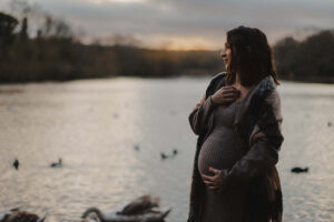 Image of a pregannt mother, standing in front of a lake in London. She is smiling and has one hand on her belly and one hand on her necklace.