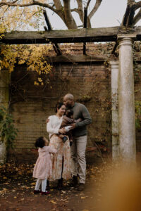Family of 4 embracing, dad, mum, baby and little girl, kissing and hugging in Hampstead Heath The Pergola during their autumn Family Photoshoot in London.