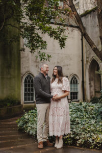 Maternity Photoshoot in St Dunstan in the East Church. Man and Woman standing next to tree in church ruins, smiling at eachother. The man is touching his wife baby bump.