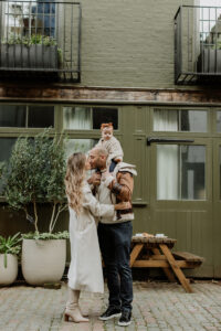 Family Photoshoot in St Luke's Mews, Notting Hill during winter. Dad and mum are kissing while their little girl is on the dad's shoulders.