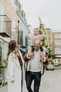 Family Photoshoot in St Luke's Mews, Notting Hill during their winter family photos. Dad and mum are touching while their little girl is on the dad's shoulders having fun.